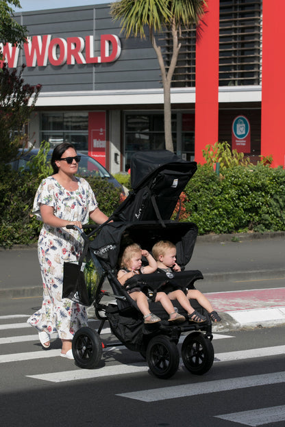 A woman pushes a three seat pushchair with a KinderPod double buggy and a baby recliner seat attached on the canopy with three young children seated in across a crosswalk in front of a shopping center building. 
