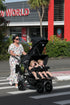 A woman pushes a three seat pushchair with a KinderPod double buggy and a baby recliner seat attached on the canopy with three young children seated in across a crosswalk in front of a shopping center building. 