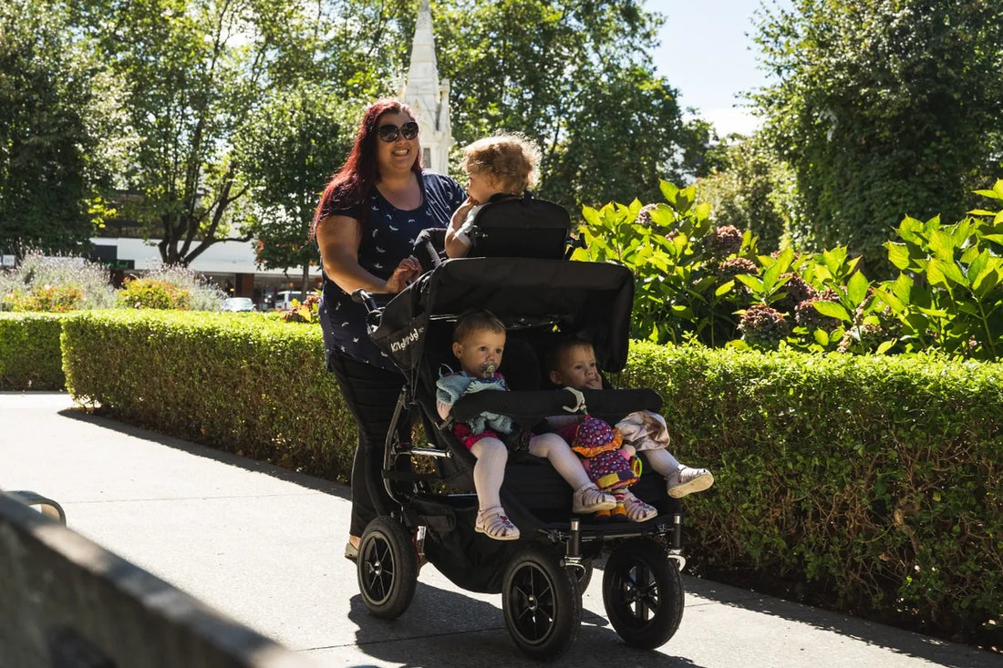 Childminder walking three young children in a KinderPod multi-seat pushchair on a park pathway.