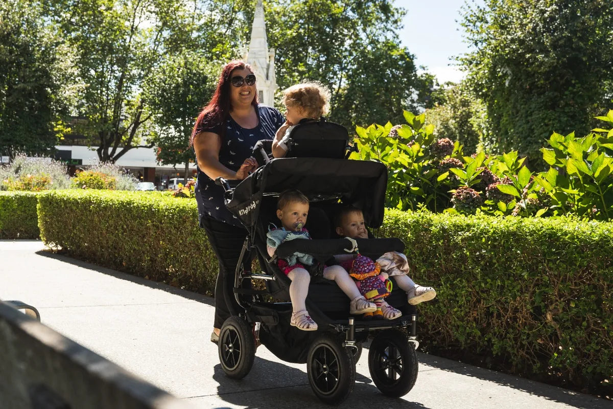 Childminder walking three young children in a KinderPod multi-seat pushchair on a park pathway.