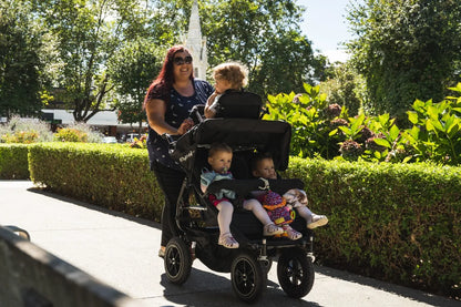Childminder walking three young children in a KinderPod multi-seat pushchair on a park pathway.