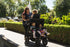 Childminder walking three young children in a KinderPod multi-seat pushchair on a park pathway.